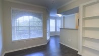 This single-floor residence showcases a well-designed open layout that maximizes both space and natural light. The main living area features rich, dark hardwood flooring that extends throughout, providing a striking contrast against the neutral-toned walls. Elegant crown molding frames the ceilings, adding a refined architectural detail to the space.

Large windows fitted with horizontal blinds flood the living area with generous amounts of natural light, creating a bright and airy atmosphere. A standout feature is the bay window nook, which curves gracefully and offers a cozy, sunlit alcove — ideal for a reading corner or casual seating. A glass-paneled entry door further enhances the sense of openness.

Adjacent to the main living area, a pass-through counter with a granite or stone surface connects to the kitchen, providing both functional workspace and a visual separation between the two areas. Light wood cabinetry is visible above the counter, contributing to a warm aesthetic. A pendant light fixture above the pass-through adds a subtle design accent. Overall, the space presents a clean, classic interior with practical architectural details throughout.