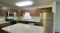 This single-floor space is thoughtfully designed to offer both functionality and comfort. The kitchen serves as a standout feature, showcasing rich, warm-toned wood cabinetry that provides generous storage both above and below the countertops. Smooth granite countertops with a classic speckled finish extend across the workspace, including a spacious island or peninsula that adds additional prep and seating space, enhancing the kitchen's practical appeal. Stainless steel appliances, including a refrigerator, dishwasher, range, and built-in microwave, lend the kitchen a sleek, modern aesthetic. A double basin sink is neatly positioned along the countertop run, complemented by a chrome faucet. Bright flush-mount ceiling lighting illuminates the kitchen effectively, creating a clean and inviting atmosphere throughout. The neutral wall tones and light-colored surfaces contribute to an open, airy feel, making the space feel welcoming and easy to maintain. Overall, the space is designed with practicality in mind, offering a well-equipped kitchen layout that flows naturally and supports everyday living.
