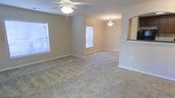 This single-floor residence offers a welcoming and functional layout centered around an open-concept living and dining area. The living room is generously sized, featuring neutral-toned carpet flooring and warm beige walls that create a bright, comfortable atmosphere. A ceiling fan with an integrated light fixture provides both air circulation and ambient lighting, while multiple windows fitted with blinds allow for ample natural light throughout the day.

The space flows seamlessly into a dedicated dining area, distinguished by an elegant pendant light fixture that adds a touch of classic charm. The transition between the living and dining spaces is open and unobstructed, lending a sense of spaciousness to the overall floor plan.

A pass-through window connects the main living area to the kitchen, offering convenience and visual connectivity between the spaces. The kitchen area, partially visible through the pass-through, features a contrasting countertop that adds a subtle design detail. Clean white baseboards trim the perimeter of the rooms, complementing the neutral color palette throughout.

Overall, the space presents a clean, move-in-ready interior with a practical and open layout suited for everyday living and entertaining.