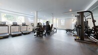 Located on Santa Monica Boulevard in Los Angeles, this well-appointed fitness facility offers a generously sized, open-plan workout space designed to support a full range of training activities. The interior is finished with a clean, contemporary aesthetic — crisp white walls, recessed lighting, and a durable rubberized floor that spans the entire area.

Large floor-to-ceiling windows and multiple smaller panes along the perimeter flood the space with natural light while providing views of the surrounding urban streetscape. Structural white columns punctuate the floor plan, adding architectural definition without interrupting the open layout.

The strength training area is comprehensively equipped, featuring a squat rack with weight plates, cable machines, adjustable benches, and a well-organized storage rack stocked with kettlebells, medicine balls, and stability balls. A mirrored wall enhances the sense of depth in this zone. The cardio section includes treadmills, a stationary bike, and stair climbers, all positioned near the windows to take advantage of natural light. A water fountain is conveniently installed along one wall. The space is branded with a stylish neon sign, lending a polished, boutique-gym feel. Overall, the facility combines functionality and modern design in a prime West Los Angeles location.
