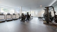 Located on Santa Monica Boulevard in Los Angeles, this well-appointed fitness facility offers a generously sized, open-plan workout space designed to support a full range of training activities. The interior is finished with a clean, contemporary aesthetic — crisp white walls, recessed lighting, and a durable rubberized floor that spans the entire area.

Large floor-to-ceiling windows and multiple smaller panes along the perimeter flood the space with natural light while providing views of the surrounding urban streetscape. Structural white columns punctuate the floor plan, adding architectural definition without interrupting the open layout.

The strength training area is comprehensively equipped, featuring a squat rack with weight plates, cable machines, adjustable benches, and a well-organized storage rack stocked with kettlebells, medicine balls, and stability balls. A mirrored wall enhances the sense of depth in this zone. The cardio section includes treadmills, a stationary bike, and stair climbers, all positioned near the windows to take advantage of natural light. A water fountain is conveniently installed along one wall. The space is branded with a stylish neon sign, lending a polished, boutique-gym feel. Overall, the facility combines functionality and modern design in a prime West Los Angeles location.