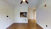 This single-floor residence showcases an open and airy layout anchored by warm hardwood flooring that extends throughout the main living areas. The living space is appointed with a ceiling fan, adding both functionality and a subtle decorative touch to the bright, white-walled interior.

A defining architectural feature is the pass-through kitchen, framed by a partial wall with a granite or stone-topped counter that serves as both a practical workspace and an informal bar or serving area. The kitchen is equipped with stainless steel appliances, including a microwave and range, set against dark cabinetry and a tiled backsplash, creating a pleasing contrast with the lighter surroundings.

A hallway, illuminated by a modern track lighting fixture mounted to the ceiling, leads further into the residence, suggesting additional rooms beyond the main living area. Clean white walls and crisp baseboards contribute to an overall sense of tidiness and simplicity. The open floor plan between the living area and kitchen promotes a connected, functional flow, making the space well-suited for everyday living and casual entertaining.