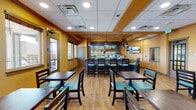 Located at 22000 E Quincy Ave in Aurora, Colorado, this single-floor bar and dining venue offers a warm, casual atmosphere designed for comfortable social gatherings. The space is defined by its rich amber-painted walls, light hardwood flooring, and a drop ceiling with recessed lighting that creates an inviting, well-lit environment throughout.

The centerpiece of the space is a fully outfitted bar featuring a stainless steel counter, multiple draft beer taps, an ice machine, glass-front refrigerators stocked with beverages, and backlit glass shelving displaying an extensive collection of glassware. Pendant lights with dark industrial-style shades hang above the bar, adding a stylish focal point. Wall-mounted flat-screen televisions are positioned throughout the venue for easy viewing from most seating areas.

Dining seating is generously distributed across the open floor plan, featuring dark wood tables paired with chairs and banquette benches upholstered in teal cushions and striped fabric. A cozy banquette section along one wall is adorned with framed vintage-style wine and entertainment posters, lending a lively, curated character to the space. Multiple exterior-facing windows and glass-panel doors allow natural light to filter in, providing views of the surrounding area. The layout accommodates both intimate dining and larger group settings with ease.