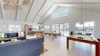 Located at 26 Mullein Dr in Hampstead, NC, this single-floor community space is designed to impress with its dramatic vaulted ceilings accented by exposed white beams that soar overhead, flooding the open-concept layout with an airy, expansive feel. Light hardwood-style flooring runs throughout, tying together the living, dining, and kitchen areas seamlessly.

The living room anchors the space with a striking wood-clad accent wall featuring a mounted flat-screen TV, complemented by stylish blue sectional sofas, warm leather accent chairs, and woven pendant light fixtures that add a coastal touch. A pool table provides an additional recreational focal point.

The dining area accommodates a large gathering table with wicker-style chairs, positioned adjacent to floor-to-ceiling sliding glass doors that open to an outdoor pool area, allowing abundant natural light and views of the surrounding community. The kitchenette features gray cabinetry, a long white quartz-topped island with bar seating, and a retro-style refrigerator, making it ideal for hosting.

A dedicated office features whitewashed brick-textured walls, a round conference table with upholstered chairs, built-in shelving, and large windows offering natural light — perfect for focused work or small meetings. Together, these spaces create a versatile, welcoming environment suited to both social gatherings and productive work.