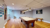 Located at 2881 Richmond Rd in Ottawa, this billiards room is a well-appointed recreational space designed for leisure and social activity. The room centers around a full-size pool table with a rich cherry wood finish, positioned beneath a classic brass chain-hung pendant light fixture with multiple shades that provide focused, even illumination over the playing surface. The flooring throughout is warm-toned hardwood, complementing the neutral grey walls and white baseboards for a clean, polished look. A drop tile ceiling with recessed pot lights ensures bright, uniform lighting across the entire space. Seating is provided by a wooden bench along the wall, and a wall-mounted cue rack with a full set of billiard balls and cues keeps equipment neatly organized and accessible. Decorative landscape artworks adorn the walls, adding visual interest and a relaxed atmosphere. Large windows along one wall allow natural light to filter in, offering views of surrounding greenery. A full-height glass partition wall on one side opens the room visually to an adjacent corridor and amenity area, enhancing the sense of openness. Baseboard heating runs along the perimeter, ensuring year-round comfort. The room is accessed via a standard door and is well-suited as a shared amenity space.