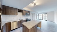 This well-appointed two-bedroom, two-bathroom residence offers a thoughtfully designed open-concept layout finished with contemporary materials throughout. The kitchen is a standout feature, showcasing rich dark wood cabinetry accented by an eye-catching herringbone tile backsplash, stainless steel appliances including a dishwasher, range, and microwave, and a generous marble-topped island on casters that adds both prep space and visual elegance. Light hardwood flooring flows through the main living and kitchen areas, complementing the clean, neutral palette.

The open living area is bathed in natural light through expansive floor-to-ceiling windows and sliding glass doors that frame striking urban skyline views, creating a bright and airy atmosphere. The primary bedroom is a well-sized retreat with plush carpeting and large windows overlooking the cityscape. Its en-suite bathroom features a floating double-vanity with a crisp white quartz countertop, dual undermount sinks, wood-toned cabinetry, and a large mirror. A dedicated walk-in closet provides excellent storage. The second bedroom, also carpeted, benefits from its own large window with urban views and convenient access to the second full bathroom. Together, these spaces deliver a modern, functional, and stylish urban living experience.