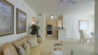 This single-story residence is designed with an open-concept layout that seamlessly connects the main living area to the kitchen. The living room features vaulted ceilings fitted with a ceiling fan, contributing to a spacious and airy atmosphere. Light-toned carpet flows throughout the living space, complementing the neutral cream and white color palette that defines the interior. A large framed artwork adorns one wall, while a lush indoor plant adds a natural accent to the corner of the room. The living area is centered around a comfortable sofa with decorative cushions and a low coffee table.

The kitchen is visible through an open passageway and features light wood cabinetry, a white electric range, a built-in microwave, and a compact peninsula that serves as a casual dining or prep surface. Tile flooring in the kitchen provides a practical contrast to the carpeted living space. Natural light filters in through a window, brightening the kitchen area. An arched doorway detail adds an architectural touch to the interior. The front entry door is positioned to the right, and the overall layout promotes a functional and comfortable flow throughout the home.