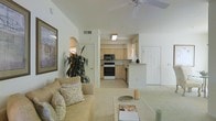 This single-story residence is designed with an open-concept layout that seamlessly connects the main living area to the kitchen. The living room features vaulted ceilings fitted with a ceiling fan, contributing to a spacious and airy atmosphere. Light-toned carpet flows throughout the living space, complementing the neutral cream and white color palette that defines the interior. A large framed artwork adorns one wall, while a lush indoor plant adds a natural accent to the corner of the room. The living area is centered around a comfortable sofa with decorative cushions and a low coffee table.

The kitchen is visible through an open passageway and features light wood cabinetry, a white electric range, a built-in microwave, and a compact peninsula that serves as a casual dining or prep surface. Tile flooring in the kitchen provides a practical contrast to the carpeted living space. Natural light filters in through a window, brightening the kitchen area. An arched doorway detail adds an architectural touch to the interior. The front entry door is positioned to the right, and the overall layout promotes a functional and comfortable flow throughout the home.