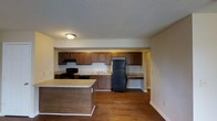 This single-floor space is designed with functionality and practicality in mind. The kitchen serves as the focal point of the layout, showcasing rich, dark wood cabinetry that lines the full width of the back wall, offering generous storage and a warm, traditional aesthetic. Black appliances, including a freestanding range and a full-size refrigerator, complement the cabinetry and add a cohesive, classic look. A double sink is centrally positioned along the counter, flanked by ample workspace with light-toned countertops. A built-in desk or utility nook is tucked to the side, providing additional functionality. The kitchen island extends toward the living area, creating a natural division of space while offering extra counter space for meal preparation or casual dining. Rich hardwood flooring runs throughout, adding warmth and depth to the interior. The textured ceiling and flush-mount lighting keep the design grounded and practical. The open-concept flow between the kitchen and adjacent living areas enhances the sense of space, making the layout feel connected and easy to navigate. Overall, the space offers a straightforward, well-organized design suited for everyday living.