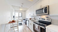 Located at 333 University Ave in Kingston, Ontario, this contemporary one-bedroom, one-bathroom unit offers a well-designed, efficient layout spread across a single floor. The open-concept living and kitchen area flows seamlessly, making excellent use of the available space. The kitchen is outfitted with sleek white cabinetry, a white subway tile backsplash, stainless steel appliances including an electric range, over-the-range microwave, dishwasher, and refrigerator, along with light wood-tone flooring that extends throughout the unit. A compact dining area with a white table and chairs sits adjacent to the kitchen, ideal for casual meals. The living room is bright and airy, featuring exposed concrete ceiling details that add an industrial character, and a large window framing expansive views of the Kingston skyline and surrounding landscape. The bedroom is a quiet, comfortable retreat with its own large window offering sweeping outdoor views, light hardwood flooring, and a built-in closet with organizers. White walls and abundant natural light throughout the unit contribute to a clean, modern ambiance. A hallway connects the main living areas to the bathroom, completing this thoughtfully laid-out urban residence.