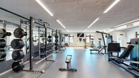 Located at 4545 Laclede Ave in St. Louis, Missouri, this well-appointed fitness center offers a comprehensive workout facility across a single, thoughtfully organized floor. The space is illuminated by long linear LED fixtures set into an acoustic tile ceiling, creating a bright and energizing environment throughout.

The facility is divided into distinct workout zones. A dedicated strength training area features a full power rack, a complete dumbbell rack stocked with a wide range of free weights, and a flat bench, all positioned in front of large floor-to-ceiling mirrors that enhance the sense of space. A separate cardio section houses multiple treadmills and a cable machine, each accompanied by wall-mounted televisions, keeping users entertained during workouts.

Additional equipment includes a cable crossover machine, elliptical trainer, rowing machine, and a leg press unit, ensuring users have access to both upper- and lower-body training options. A distinct functional training corner is accented by a bold, patterned red and white wallcovering, adding a vibrant design element to the space. This zone includes wall-mounted storage for resistance bands and medicine balls, as well as a barbell storage rack.

Natural light filters in through multiple wood-framed windows along the perimeter walls, offering views of the surrounding neighborhood. The flooring transitions between durable rubber gym flooring and hardwood-style surfaces across zones, providing both safety and aesthetic appeal.