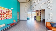 Located at 20080 West Dixie Highway in Miami, FL, this striking single-floor lobby space makes a bold architectural statement from the moment you enter. The space is defined by its dramatically high ceilings, which amplify the sense of openness and grandeur throughout. A standout feature is the cascading, lattice-like sculptural wall installation in white, which climbs from floor to ceiling and serves as a captivating artistic focal point.

Vibrant teal accent walls energize the interior, complemented by an eclectic mix of seating arrangements. These include a bold teal upholstered bench, a curved orange sofa adorned with colorful throw pillows, sleek modern benches along the teal walls, and a yellow-green reception or lounge desk. The variety of seating zones creates distinct areas for gathering, waiting, or socializing within the open floor plan.

Original works of art are displayed throughout, adding a gallery-like quality to the space. Large floor-to-ceiling windows and glazed entry doors flood the interior with abundant natural light and offer views of the outdoors, enhancing the airy, welcoming atmosphere.

The flooring features wood-look tile laid in a clean, linear pattern that unifies the entire space. Together, the bold color palette, artistic installations, and thoughtful design create a lively, contemporary environment reflective of Miami's vibrant aesthetic.