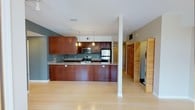 This single-floor space is designed with an open-concept layout that promotes a seamless flow between living and kitchen areas. The kitchen serves as the heart of the space, showcasing rich, warm-toned wood cabinetry that extends from base to upper levels, providing generous storage. A peninsula-style counter with a smooth light-colored surface extends outward, creating a natural division between the kitchen and the main living area while offering additional workspace and seating potential. Stainless steel appliances, including a refrigerator, range, and microwave, lend a contemporary, polished feel to the kitchen. Pendant lighting hangs above the counter, adding focused illumination and a stylish accent. The flooring throughout is light-toned hardwood, contributing to a bright and airy atmosphere. Structural columns are cleanly finished in white, integrating naturally into the open design. A set of louvered bi-fold doors on one side conceals a laundry area with a stacked washer and dryer, keeping functional utilities neatly tucked away. The walls feature a subtle blue-gray paint that complements the warm wood tones, and recessed and track lighting in the ceiling ensures the entire space is well-lit. Overall, the space presents a clean, modern aesthetic with thoughtful functionality.
