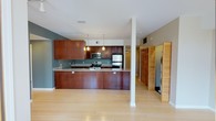 This single-floor space is designed with an open-concept layout that promotes a seamless flow between living and kitchen areas. The kitchen serves as the heart of the space, showcasing rich, warm-toned wood cabinetry that extends from base to upper levels, providing generous storage. A peninsula-style counter with a smooth light-colored surface extends outward, creating a natural division between the kitchen and the main living area while offering additional workspace and seating potential. Stainless steel appliances, including a refrigerator, range, and microwave, lend a contemporary, polished feel to the kitchen. Pendant lighting hangs above the counter, adding focused illumination and a stylish accent. The flooring throughout is light-toned hardwood, contributing to a bright and airy atmosphere. Structural columns are cleanly finished in white, integrating naturally into the open design. A set of louvered bi-fold doors on one side conceals a laundry area with a stacked washer and dryer, keeping functional utilities neatly tucked away. The walls feature a subtle blue-gray paint that complements the warm wood tones, and recessed and track lighting in the ceiling ensures the entire space is well-lit. Overall, the space presents a clean, modern aesthetic with thoughtful functionality.