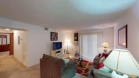 Located at 1305 Harrison Circle in Pell City, Alabama, this single-floor home offers a well-organized layout designed for comfortable everyday living. The living room serves as the heart of the home, featuring plush carpeting, neutral white walls, and a large sliding glass door with vertical blinds that allows generous natural light to fill the space. The open layout connects seamlessly to the dining area, which is positioned adjacent to the kitchen and features a round glass-top table with wrought-iron chairs beneath a flush-mount ceiling light. The kitchen is equipped with white cabinetry, a full-size refrigerator, microwave, and a sink, with light tile flooring and a pass-through counter opening to the dining space. Two bedrooms provide restful retreats, both carpeted and featuring textured ceilings, closet storage, and windows dressed with dark curtains. The primary bedroom offers dual closets and ample floor space, while the second bedroom provides a cozy, well-proportioned area. The bathroom includes a single-sink vanity with under-cabinet storage, tile flooring, a toilet, and a shower with a decorative curtain. Throughout the home, a consistent use of light tones and carpet creates a cohesive and welcoming atmosphere.