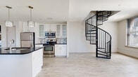 Located in the heart of Cleveland's vibrant East Sixth Street corridor, this two-floor residence offers a thoughtfully designed layout with modern finishes throughout. The main level centers around an open-concept kitchen outfitted with white shaker-style cabinetry featuring glass-front uppers, dark granite countertops, stainless steel appliances, and a mosaic tile backsplash. A large island with pendant lighting provides additional prep and seating space, while a stacked washer and dryer unit is conveniently integrated nearby. Light wood-look flooring flows seamlessly throughout both levels, enhancing the bright, airy feel of the space. A striking black spiral staircase serves as both a functional and architectural centerpiece, connecting the two floors with visual drama. The upper level opens into a generously sized living area bathed in natural light from multiple arched windows that frame dynamic views of the Cleveland skyline. Double-height ceilings in portions of the space amplify the sense of openness. The bathroom features large-format tile flooring, a glass-enclosed walk-in shower, and an adjacent walk-in closet with wire shelving. Additional rooms offer flexible use, and a utility hallway provides access to the electrical panel and smart thermostat.