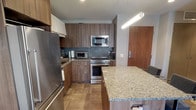 This contemporary single-floor residence is thoughtfully designed with a cohesive palette of warm neutrals, rich wood tones, and polished stainless steel accents. The kitchen is a standout, outfitted with flat-front walnut-finish cabinetry, granite countertops, a textured tile backsplash, and a full suite of stainless steel appliances including a French door refrigerator, electric range, and over-the-range microwave. A granite-topped island with bar seating provides an ideal space for casual dining and entertaining, illuminated by a sleek linear pendant light. The living area offers a comfortable, intimate atmosphere with warm grey walls, recessed lighting, and curated wall art that adds a gallery-like character to the space. The bedroom is a serene retreat featuring neutral-toned walls, plush carpeting, recessed lighting, and a decorative shoji-style shelving unit that adds an elegant architectural detail. Multiple windows allow natural light to filter in gently. The bathroom is finished to a high standard, with large-format stone-look floor and wall tiles, a walk-in glass shower with a mosaic tile niche, a granite vanity, and backlit mirrors, creating a spa-inspired atmosphere throughout.
