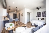 This single-floor unit showcases a contemporary, open-concept design that seamlessly integrates the living, dining, and kitchen areas. Warm hardwood-style flooring flows throughout, complementing the clean white walls and recessed lighting. An eye-catching industrial touch is introduced by exposed silver ductwork running along the ceiling in the main living and kitchen zones, adding an urban loft character to the otherwise modern aesthetic.

The kitchen is well-appointed with stainless steel appliances, including a full-size refrigerator, range, microwave, and dishwasher. Two-tone cabinetry pairs dark lower cabinets with white upper cabinets, topped with light countertops. A breakfast bar with seating for two extends into the living area, making it ideal for casual dining.

The living space accommodates a comfortable seating arrangement, while the bedroom area is defined by a sliding closet, a ceiling fan, and a large floor-to-ceiling window with balcony access, offering outdoor views and abundant natural light. The bathroom features a streamlined vanity with dark cabinetry, matte black fixtures, and a stacked in-unit washer and dryer, adding practical convenience to this well-designed space.