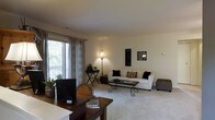 This single-floor space presents a thoughtfully arranged living area anchored by neutral-toned, carpet flooring that extends throughout the room, contributing to a warm and cohesive atmosphere. A large sliding glass door along one wall allows generous natural light to filter in through sheer curtains, offering a view of an outdoor balcony area framed by lush greenery. The living room is comfortably appointed with a white sofa accented by decorative pillows and complemented by a sculptural iron and glass coffee table, creating an elegant focal point. A tall floor lamp provides warm ambient lighting, while two decorative wall panels add visual interest to the neutral walls. Adjacent to the living area, a defined workspace features a rich dark wood desk paired with a wicker chair, accommodating both productivity and comfort. A matching dark wood side table with a slim profile adds further functionality near the window. Potted plants placed throughout the space introduce natural texture and life. The overall ambiance is calm, clean, and inviting, with a balanced mix of modern and transitional design elements.