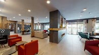 This thoughtfully designed single-floor amenity space seamlessly blends industrial, rustic, and contemporary elements throughout its various distinct zones. Reclaimed wood accent walls, exposed brick, and corrugated metal panels create a warm yet edgy aesthetic, complemented by light-toned tile flooring that runs throughout the space.

The main lounge area serves as the centerpiece, featuring a striking dark metal double-sided fireplace framed with natural wood accents, comfortable sectional seating, and vibrant orange armchairs that add bold pops of color. Floor-to-ceiling sliding glass doors flood the space with natural light and provide views of an outdoor patio area.

A dedicated media lounge offers leather sofas, teal accent seating, and a large wall-mounted flat-screen television set against an exposed brick and reclaimed wood feature wall. Adjacent seating nooks with built-in banquette benches and eclectic ottomans provide intimate gathering spots throughout.

The game room is a standout feature, showcasing a full-size billiards table and shuffleboard set against dramatic corrugated metal walls adorned with vibrant original artwork. A separate game table area with a large-format Scrabble board adds interactive entertainment options.

A bar-height counter with black stools, a herringbone tile accent wall, and a colorful mosaic-backed bench near the outdoor access point round out the space, while curated artwork displayed throughout adds a gallery-like quality to the overall environment.
