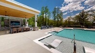 Located at 223 Long Shoals Rd in Asheville, North Carolina, this outdoor amenity space offers a thoughtfully designed resort-style pool area that serves as a premier destination for relaxation and recreation. The centerpiece is a generously sized swimming pool with crystal-clear blue water, complemented by a shallow sun shelf outfitted with contemporary lounge chairs that allow residents to recline partially submerged. An adjacent spa provides a more intimate setting for unwinding. The expansive pool deck is finished in light-toned concrete and is lined with multiple chaise lounges and seating clusters in navy blue, offering ample space for sunbathing and socializing. A covered cabana with a wood-paneled ceiling, white columns, a ceiling fan, and a stone-accented outdoor kitchen or bar area provides shaded respite and an ideal gathering spot. The entire area is enclosed by a black iron fence, with a backdrop of mature, lush trees that lend a sense of privacy and natural beauty. The multi-story residential building, clad in blue and gray siding with stone accents and private balconies, frames the courtyard on one side, creating an intimate and community-centered atmosphere. Under open Asheville skies, this outdoor space delivers a refined, amenity-rich experience.