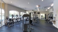 This fitness center offers a comprehensive workout environment designed to accommodate a variety of exercise routines. The space is generously sized, with a open floor plan that allows for easy navigation between equipment zones. Large arched windows line the walls, flooding the interior with abundant natural light and providing views of the surrounding greenery, creating an inviting and energizing atmosphere.

The cardio section features a row of elliptical machines and treadmills positioned along the windowed wall, making workouts feel open and connected to the outdoors. The strength training area is well-appointed with a multi-station cable machine, weight benches, and a selection of additional resistance equipment, catering to both beginner and experienced gym-goers.

Ceiling fans are distributed throughout the space to maintain comfortable airflow during intense workouts, complemented by recessed lighting that ensures consistent illumination across the entire floor. A wall-mounted television is also present, adding an entertainment option for those on cardio equipment. The flooring consists of durable, dark rubberized material, providing safety and support underfoot. The overall ambiance is clean, modern, and functional, reflecting a space purpose-built for health and fitness.