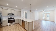 Located at 9000 Palm Passage Loop in Summerville, SC, this well-appointed 2-bedroom, 2-bathroom single-floor home offers a clean, contemporary design with thoughtful finishes throughout. The open-concept kitchen and living area serves as the heart of the home, where a large center island with a light granite countertop and undermount sink takes center stage, illuminated by a trio of polished pendant lights. The kitchen features gray shaker-style cabinetry, a herringbone tile backsplash, stainless steel appliances including a French door refrigerator, range, microwave, and dishwasher, and a built-in wine rack. Light hardwood-style flooring runs seamlessly through the entire space. The living room is bright and airy, highlighted by a ceiling fan, recessed lighting, and glass doors that open to an outdoor balcony with natural views. Both bedrooms are spacious and well-lit with natural light from large windows. The primary bathroom offers a dual-sink vanity with granite counters, gray cabinetry, and a large framed mirror. A dedicated laundry room houses a full-size top-load washer and dryer with overhead wire shelving for added storage. The overall ambiance is fresh, modern, and move-in ready.