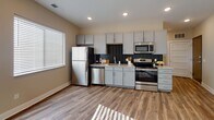 This single-floor space features a thoughtfully designed open layout that seamlessly connects the living area and kitchen, creating a bright and cohesive environment. Warm-toned wood-look flooring extends throughout the main living areas, complemented by recessed lighting and neutral-colored walls that enhance the sense of openness. Large windows fitted with blinds allow generous natural light to fill the space, casting distinctive light patterns across the floors.

The kitchen is well-appointed with gray shaker-style cabinetry, granite-look countertops, and a full suite of stainless steel appliances including a refrigerator, electric range, microwave, and dishwasher. A dark tile backsplash provides a refined contrast to the lighter cabinetry, adding visual depth to the cooking area.

The bathroom features a gray vanity with a granite countertop, an integrated sink, a large mirror, and a shower-tub combination enclosed by a neutral striped curtain. Wood-look flooring continues into this space, maintaining design consistency throughout.

One bedroom is finished with plush carpet flooring and neutral walls, offering a comfortable retreat. A dedicated walk-in closet with a full-width hanging rod and additional shelving provides practical storage. The overall design is clean, modern, and move-in ready.