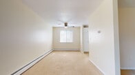 This single-floor unit offers a straightforward, functional layout comprising a living area, bedroom spaces, a kitchen, and a full bathroom. The living area is generously sized, featuring neutral-toned wall-to-wall carpeting, cream-colored walls, and a textured ceiling with a ceiling fan and integrated light fixture. A window along the far wall allows natural light to filter in, while a wall-mounted air conditioning unit and baseboard heating provide year-round climate control.

The bedroom areas mirror the living space in finish, with plush carpeting, baseboard heating, and windows dressed with horizontal blinds. One bedroom includes a spacious reach-in closet with a hanging rod, offering practical storage. The layout connects rooms through a straightforward hallway.

The kitchen features white cabinetry, dark laminate countertops, and tile flooring. An electric range with an overhead ventilation hood is included, and ample cabinet storage lines both the upper and lower walls.

The bathroom is finished with tile flooring and tile wainscoting, and includes a single-sink vanity with warm wood cabinetry, a mirror cabinet, and a combined tub and shower. A window with blinds provides ventilation and natural light. Baseboard heating is present here as well, contributing to consistent warmth throughout the unit.