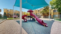 Located at 7834 Viscount Blvd in El Paso, Texas, this outdoor playground area serves as a central recreational amenity within a multi-building residential community. The space is thoughtfully designed to provide a safe and engaging environment for children, set against the backdrop of warm, tan-colored stucco buildings with teal accents that define the community's Southwestern architectural style.

At the heart of the playground stands a colorful multi-level play structure featuring bold red tube slides, a climbing tower with yellow railings and ladder rungs, and multiple access points for children of varying ages and abilities. The play equipment is installed over a cushioned rubber safety surface, providing impact absorption and a clearly defined play zone. A large teal shade sail canopy stretches overhead, offering substantial protection from El Paso's intense sun and making the space usable throughout the day.

Surrounding the core play area is a generous expanse of compacted gravel, bordered by stone edging and metal fencing that secures the perimeter. Mature trees are scattered throughout and adjacent areas, contributing natural shade and greenery. Well-maintained grass borders and landscaped pathways connect the playground to the broader residential grounds. The overall ambiance is family-friendly and community-oriented, offering a practical and welcoming outdoor space designed for active play and outdoor enjoyment.