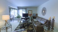 This single-floor residence offers a thoughtfully arranged layout that balances comfort and functionality. The living room serves as the heart of the home, featuring light-colored carpeting that adds warmth underfoot and soft, neutral walls that create a calm, welcoming atmosphere. Large windows dressed with decorative valances allow natural light to fill the space, illuminating a generous seating area anchored by a plush leather sofa and a stylish glass-topped coffee table resting on a boldly patterned area rug. Decorative accents, including a sunburst wall mirror and lush indoor plants, add character and life to the room. A dedicated media and entertainment setup is neatly organized along one wall, complementing the overall livability of the space. The dining area is equally inviting, featuring a round glass-top table paired with elegant wrought-iron chairs with cushioned seats, ideal for intimate gatherings. A ceiling fan overhead provides both light and airflow, while large windows offer an abundance of natural light and outdoor views. Framed artwork adorns the walls, contributing to the space's refined yet approachable aesthetic throughout.