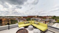 This rooftop terrace offers an exceptional outdoor living experience elevated above the surrounding urban landscape. The space is designed for relaxation and social gathering, centered around a generously sized sectional sofa dressed in bold chartreuse cushions and accented with colorful patterned throw pillows. The seating arrangement is complemented by woven pouf ottomans that add a casual, textural element to the setting. A substantial fire pit serves as a natural focal point, providing warmth and ambiance during cooler evenings. The terrace is finished with clean, light-toned tile flooring and enclosed by sleek glass-panel railings that preserve unobstructed sightlines in every direction. From this elevated vantage point, sweeping panoramic views of the city stretch across the horizon, showcasing a mix of brick mid-rise buildings, commercial corridors, and open sky. The open-air design invites abundant natural light throughout the day, while the dramatic cloudscape overhead enhances the sense of being above the city. This rooftop space strikes a balance between modern, sophisticated design and inviting comfort, making it an ideal venue for both quiet retreats and lively outdoor gatherings.