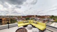 This rooftop terrace offers an exceptional outdoor living experience elevated above the surrounding urban landscape. The space is designed for relaxation and social gathering, centered around a generously sized sectional sofa dressed in bold chartreuse cushions and accented with colorful patterned throw pillows. The seating arrangement is complemented by woven pouf ottomans that add a casual, textural element to the setting. A substantial fire pit serves as a natural focal point, providing warmth and ambiance during cooler evenings. The terrace is finished with clean, light-toned tile flooring and enclosed by sleek glass-panel railings that preserve unobstructed sightlines in every direction. From this elevated vantage point, sweeping panoramic views of the city stretch across the horizon, showcasing a mix of brick mid-rise buildings, commercial corridors, and open sky. The open-air design invites abundant natural light throughout the day, while the dramatic cloudscape overhead enhances the sense of being above the city. This rooftop space strikes a balance between modern, sophisticated design and inviting comfort, making it an ideal venue for both quiet retreats and lively outdoor gatherings.