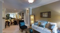 This thoughtfully designed single-floor residence showcases an open-concept layout that seamlessly connects the living and dining areas, creating a cohesive and inviting atmosphere. The living area is anchored by a comfortable white sofa dressed with teal and striped accent pillows, complemented by a sleek black accent chair adorned with a bold turquoise cushion. A low-profile coffee table grounds the seating arrangement, while a distinctive illuminated decorative column with a warm amber glow serves as a striking focal point that adds artistic character to the space. Crown molding runs along the ceiling line, lending a refined architectural detail throughout. Soft, neutral wall tones in warm taupe provide a sophisticated backdrop that highlights the carefully curated furnishings. Recessed lighting and a flush-mount ceiling fixture ensure the space is well-lit and welcoming. The dining area, visible through the open layout, features a rich wood dining table surrounded by woven rattan chairs, evoking a relaxed yet elegant ambiance. Built-in shelving is visible in the background, offering functional display and storage. The overall aesthetic is contemporary and polished, with a cohesive color palette of neutrals, teal, and warm gold tones that create a harmonious and livable environment.