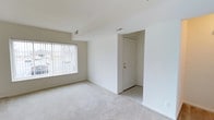 This single-floor space presents a clean, minimalist interior defined by its bright white walls and light-toned carpeted flooring. The main living area is open and airy, benefiting from a generously sized window fitted with vertical blinds that allow natural light to filter into the room while providing privacy. The window offers a view of the surrounding exterior environment. The entry area, visible from the main room, features tile flooring and a solid white entry door with gold-toned hardware, creating a clear transition zone between the interior and exterior. HVAC vents are positioned along the ceiling, ensuring climate control throughout the space. The vaulted or angled ceiling detail adds a subtle architectural element, contributing to an overall sense of openness. The neutral color palette — spanning the walls, ceiling, and flooring — creates a versatile canvas adaptable to a variety of interior styles and furnishing choices. The space is well-maintained and move-in ready, presenting a straightforward and functional layout suited for everyday living.