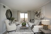 This single-floor space presents a thoughtfully designed living area that exudes a calm, sophisticated ambiance through its carefully curated neutral color palette. The room is anchored by a plush white sofa ensemble complemented by coordinating armchairs, all arranged around a wooden coffee table atop a textured area rug in soft grey tones. A striking large-scale abstract artwork in muted blues and greys serves as a dramatic focal point on one wall, adding artistic depth to the space. Natural light filters generously into the room through a sliding glass door dressed with floor-length sheer curtains, offering access to an outdoor area and providing a bright, airy atmosphere. The walls are finished in a warm greige tone that harmonizes with the soft furnishings, while crown molding adds a refined architectural detail along the ceiling perimeter. The textured ceiling adds subtle dimension overhead. Decorative accents, including lush greenery, ceramic vessels, and layered throw pillows in complementary patterns, contribute to the cohesive and polished interior aesthetic throughout the living space.