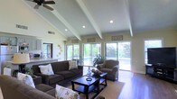 This single-floor home is designed with comfort and openness in mind, offering a welcoming living environment highlighted by striking architectural details and abundant natural light. The main living area draws immediate attention with its vaulted, beamed ceiling accented by recessed lighting and a ceiling fan, creating a sense of spaciousness and airiness throughout. Large sliding glass doors span much of the rear wall, flooding the interior with natural light and providing sweeping views of the outdoor pool area and lush surrounding greenery — seamlessly blending indoor and outdoor living. The living room is well-appointed with plush, dark-toned sofas and an armchair arranged around a bold, dark wood coffee table, all anchored by a neutral area rug over tile flooring. Decorative wall art and ornamental plate displays add personality to the soft, neutral-toned walls. A glimpse into the adjacent kitchen reveals light cabinetry and countertops, suggesting a functional and cohesive layout. The overall ambiance of the home is warm, relaxed, and inviting, with a design that balances casual comfort with tasteful style — ideal for both everyday living and entertaining.
