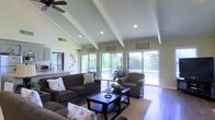 This single-floor home is designed with comfort and openness in mind, offering a welcoming living environment highlighted by striking architectural details and abundant natural light. The main living area draws immediate attention with its vaulted, beamed ceiling accented by recessed lighting and a ceiling fan, creating a sense of spaciousness and airiness throughout. Large sliding glass doors span much of the rear wall, flooding the interior with natural light and providing sweeping views of the outdoor pool area and lush surrounding greenery — seamlessly blending indoor and outdoor living. The living room is well-appointed with plush, dark-toned sofas and an armchair arranged around a bold, dark wood coffee table, all anchored by a neutral area rug over tile flooring. Decorative wall art and ornamental plate displays add personality to the soft, neutral-toned walls. A glimpse into the adjacent kitchen reveals light cabinetry and countertops, suggesting a functional and cohesive layout. The overall ambiance of the home is warm, relaxed, and inviting, with a design that balances casual comfort with tasteful style — ideal for both everyday living and entertaining.