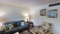 This single-floor space offers a warm and inviting atmosphere, centered around a thoughtfully arranged living area. The room features light, neutral-toned walls that create a calm and airy ambiance, complemented by plush carpeted flooring that adds a sense of warmth underfoot. The seating arrangement includes a charcoal-gray sofa accented with decorative throw pillows and a coordinating floral-patterned armchair, offering comfortable and functional lounging space. A classic table lamp on a dark end table provides soft, ambient lighting that enhances the room's cozy feel. Wall décor, including a framed birdcage art piece, adds a subtle decorative touch without overwhelming the clean, understated aesthetic. The layout is efficient and well-organized, making good use of the available space. The overall design lends itself to a relaxed, livable environment that balances simplicity with comfort. The light-colored walls and soft furnishings work together to create a welcoming setting suited for everyday living.