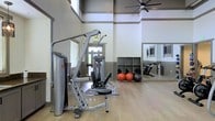 This fitness center offers a comprehensive workout environment designed to accommodate a variety of exercise routines. The space is defined by its soaring ceilings, which lend an open and airy atmosphere, complemented by large windows that invite natural light throughout. A large ceiling fan overhead ensures comfortable air circulation during workouts.

The flooring consists of smooth, hardwood-style planks that provide both durability and a clean, polished aesthetic. A multi-function strength training machine serves as a central fixture, offering users options for a wide range of resistance exercises. Along the perimeter, a built-in cubby storage unit houses an assortment of exercise balls in varying sizes, keeping the space organized and functional.

Full-length mirrors line the walls, amplifying the sense of space and allowing users to monitor their form during exercise. A stationary cycling bike is also visible, catering to cardiovascular training needs. The overall layout is thoughtfully arranged to maximize floor space, providing ample room for stretching and floor-based workouts. The combination of natural light, high ceilings, and well-organized equipment creates an inviting and motivating environment suited for a broad range of fitness activities.