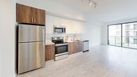 Located at 1080 NE 215th St in Miami, FL, this well-appointed one-bedroom, one-bathroom unit offers a clean, modern aesthetic with a bright and open layout. Light-toned wood-style flooring runs throughout the living spaces, complementing the crisp white walls and contributing to an airy, contemporary ambiance.

The open-concept kitchen and living area serves as the heart of the unit. The kitchen features a sleek two-tone design with white upper cabinetry and warm wood-finish lower cabinets, paired with stainless steel appliances including a refrigerator, range, microwave, and dishwasher. Light countertops and track lighting complete the polished look. The living area flows seamlessly from the kitchen and opens directly onto a private balcony through large sliding glass doors, allowing an abundance of natural light and outdoor views.

The bedroom is a well-proportioned, serene retreat with a large window framing views of the surrounding neighborhood and greenery. Three closets—including a generously sized walk-in with wire shelving—provide ample storage throughout the unit.

The bathroom is finished with large-format beige floor tiles, a wood-toned vanity with white countertop, and a standout tub-shower combination clad in bold, deep-blue glossy subway tile. An in-unit laundry area and an entrance hallway round out this thoughtfully designed Miami residence.