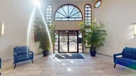 This single-floor space offers a thoughtfully designed layout spanning a generous footprint, combining an impressive entrance with a variety of functional areas. The grand lobby immediately sets the tone with soaring ceilings, a striking arched window flanked by additional transom and porthole windows, and floor-to-ceiling glass entry doors. Abundant natural light floods the polished stone tile flooring, while tall potted palms frame the entrance, adding a welcoming, resort-like ambiance.

Beyond the lobby, a comfortable lounge area features a dramatic exposed brick accent wall anchoring a wall-mounted flat-screen television and a sleek linear fireplace below. Leather seating, including a bold caramel-toned armchair and a gray sofa, surrounds a rustic wooden coffee table, creating a relaxed gathering space.

A dedicated conference or dining area accommodates a substantial live-edge wood table surrounded by leather chairs, complemented by a large framed vintage world map as a focal point. Recessed lighting and multiple windows providing outdoor views enhance the room's open, airy character.

The fully equipped kitchen features stainless steel appliances, modern gray cabinetry, white countertops, and a blue subway tile backsplash, alongside a beverage station. A clean, well-appointed restroom with a gray vanity and tile flooring rounds out the space.