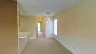 This single-floor residence offers a warm and inviting atmosphere highlighted by a clean, open layout and a cohesive neutral color palette throughout. The dining area is a standout feature, anchored by an elegant cluster pendant light fixture that adds a decorative focal point while providing warm, ambient illumination. Large windows allow natural light to fill the space, enhancing the bright and airy feel of the interior.

Soft, wall-to-wall carpeting runs throughout the main living areas, contributing to a cozy and comfortable underfoot experience. The smooth, light-toned walls and white baseboards lend a crisp, well-maintained appearance to each room. A partial wall or countertop divider separates the dining area from an adjacent kitchen space, offering a sense of openness while maintaining distinct functional zones.

A central hallway connects the living areas to the rest of the home, with a ceiling-mounted light fixture guiding the way. The hallway leads toward what appears to be additional rooms and access to the exterior, suggesting a well-organized and practical floor plan. The overall ambiance is calm, clean, and move-in ready, making it a welcoming space suited for everyday living.