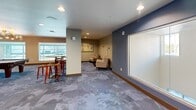 Located at 1700 Newbury Park Dr in San Jose, CA, this well-appointed community living room serves as a versatile social and recreational hub. The single-floor space is thoughtfully designed to accommodate a variety of activities and gatherings. At its centerpiece sits a full-size pool table, positioned beneath a modern sputnik chandelier that adds a stylish focal point to the room. Large windows along one wall flood the space with natural light while offering outdoor views, creating a bright and welcoming atmosphere. A high-top bar table with rustic wooden stools is positioned nearby, complemented by a vibrant large-scale abstract artwork that adds a bold pop of color to the neutral-toned walls. On the opposite end, a cozy lounge area features a plush sofa, accent armchairs, and a round coffee table arranged around a wall-mounted flat-screen TV, flanked by built-in wooden shelving units against a deep blue accent wall. Decorative wooden disc wall art adds an artistic touch to this seating nook. The patterned carpet tiles throughout give the space a contemporary, cohesive look. Double glass entry doors provide easy access, and a cue rack near the entrance reflects the room's recreational focus. The overall ambiance is modern, comfortable, and ideal for community engagement.