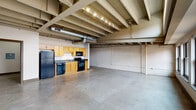 Located at 127 W Main St in the heart of downtown Louisville, Kentucky, this single-floor space embodies an industrial loft aesthetic with a range of distinctive architectural details. The open floor plan is anchored by polished concrete floors that stretch throughout, giving the space a clean, modern foundation. Exposed wooden ceiling beams and visible ductwork run overhead, adding an authentic industrial character that complements the whitewashed brick walls lining the interior.

The main living area is generously proportioned, offering a flexible, open environment bathed in natural light from a bank of large windows positioned both at eye level and clerestory height, providing views of the surrounding urban streetscape. Track lighting supplements the natural light throughout.

The kitchen is positioned along one wall and features warm honey-toned wood cabinetry, a stainless steel double sink, and a full suite of black appliances including a refrigerator, electric range, dishwasher, and over-the-range microwave, offering practical functionality within the open layout.

A private sleeping area includes a spacious closet with ample rod hanging space. The bathroom is clean and well-appointed, featuring a full tub-shower combination with white tile surround, a wood-toned vanity, and neutral finishes throughout. The overall ambiance is urban and versatile, well-suited to a variety of uses.