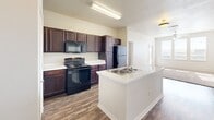 Located at 2913 Hunter Rd in San Marcos, TX, this single-floor unit offers three bedrooms and three bathrooms in a well-organized layout. The kitchen serves as a central highlight, featuring rich dark wood cabinetry extending to the ceiling for ample storage, white countertops, a center island with a built-in sink, and a full suite of black appliances including a refrigerator, electric range, and over-the-range microwave. Wood-look flooring runs throughout the kitchen and living areas, lending a cohesive and clean aesthetic. The kitchen flows openly into the living area, which is brightened by a ceiling fan and a window allowing natural light to fill the space. Each of the three bedrooms is carpeted and equipped with a ceiling fan, providing a comfortable and quiet retreat. The bedrooms are accessed via a central hallway that also connects to a dedicated closet. The bathrooms are consistent in finish, each featuring dark wood vanity cabinetry, white countertops, a tub-shower combination with white tile surround, and wood-look flooring that mirrors the rest of the unit. The overall design is clean and functional, with a neutral color palette throughout.