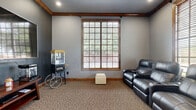 Located in Humble, TX, this single-room media lounge at 3918 Atascocita Rd is a compact yet well-appointed space designed for an engaging entertainment experience. The room is finished with neutral gray walls accented by rich wood crown molding and baseboards that lend a warm, polished character throughout.

The centerpiece of the space is a row of dark gray leather theater-style recliner seats, each equipped with built-in cup holders and control panels, providing comfortable, cinema-quality seating for multiple guests. Opposite the seating, a wall-mounted flat-screen television paired with a wooden media console creates a dedicated viewing area. Entry is provided through elegant wood-framed French doors with glass panes, offering a refined transition into the room.

Natural light filters in through multiple large windows dressed with horizontal blinds, offering both brightness and privacy control while framing outdoor greenery views. The floor is covered in a durable, textured carpet that adds warmth and sound absorption suited to a media environment. Recessed ceiling lighting and a ceiling vent ensure comfort year-round. The overall ambiance is that of a private, theater-inspired retreat designed for relaxed, immersive entertainment.