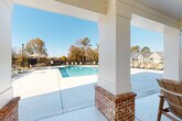 Located on Zierdt Road SW in Huntsville, Alabama, this community pool facility offers a well-appointed outdoor recreation area designed for both relaxation and social gathering. The centerpiece is a generously sized swimming pool with crystal-clear blue water, gradual entry steps, and safety railings, including an accessibility lift to accommodate all residents. The pool is enclosed within a sturdy wrought iron fence, providing security while maintaining open sightlines to the surrounding landscape and neighboring homes. A covered pavilion with white-painted columns set on brick bases offers a shaded retreat, furnished with seating for lounging out of the sun. The expansive concrete pool deck surrounds the water on all sides and is outfitted with multiple lounge chairs and umbrella-shaded tables, creating ample space for sunbathing and socializing. Adjacent to the pool, a white farmhouse-style structure with a hip roof and brick accents serves as a support building, complementing the architectural character of the broader community. Decorative lamp posts punctuate the perimeter, adding a classic touch. The open skies and mature tree backdrop visible beyond the fencing enhance the overall sense of openness and connection to the natural surroundings, making this an inviting outdoor amenity space.
