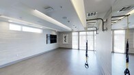 This contemporary fitness studio is thoughtfully designed to support a wide range of training activities within a clean, open floor plan. The space features warm wood-look flooring that contrasts beautifully with the crisp white walls, creating an inviting yet energizing atmosphere. A tray ceiling with integrated ambient lighting casts a soft, warm glow throughout the room, complementing the generous natural light that streams in through large, floor-to-ceiling windows dressed with horizontal blinds.

The studio is equipped with a wall-mounted TRX suspension training system and a fixed vertical ladder bar, offering versatile options for strength and functional fitness training. A pair of large flat-screen displays are mounted on one wall, ideal for guided workout sessions or instructional content. Decorative wall art adds a subtle aesthetic touch, keeping the environment visually balanced without detracting from the functional purpose of the space.

The ceiling incorporates recessed lighting, in-ceiling speakers, and HVAC vents, ensuring year-round comfort and a premium exercise environment. The overall design strikes a harmonious balance between form and function, making this studio well-suited for personal training, group fitness classes, yoga, or general exercise activities.