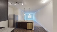 This single-floor unit offers a thoughtfully designed open-concept layout that seamlessly connects the kitchen and living area. The kitchen is a standout feature, equipped with dark espresso cabinetry, granite-look countertops, and a full suite of stainless steel appliances including a refrigerator, range, microwave, and dishwasher. A central island with a built-in sink and elegant pendant lighting serves as both a functional workspace and a natural dividing point between the kitchen and the carpeted living area. Track lighting and crown molding add a polished touch to the space.

The living area is open and bright, with neutral-toned carpet and large windows that allow natural light to filter in. Multiple bedrooms continue the warm, neutral palette with plush carpeting and windows fitted with blinds for privacy. The rooms offer generous wall space and a clean, versatile canvas.

The bathroom is finished with consistent dark cabinetry, a granite-topped vanity, a lighted mirror, and a full tub-shower combination with chrome fixtures and wood-look flooring that ties the space together cohesively. Throughout the unit, the consistent use of dark wood accents, neutral walls, and modern finishes creates a clean, contemporary aesthetic.